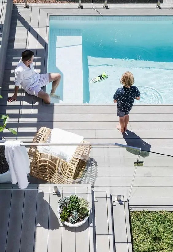 a cool modern backyard with a wooden deck, a pool, a rattan chair, some greenery and a glass fence to make the pool safe