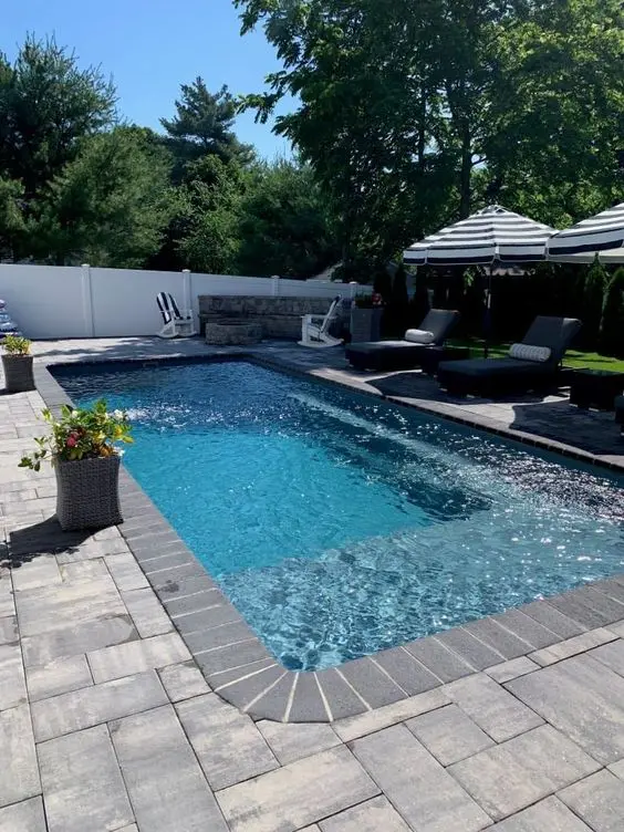 a great outdoor space with a stone deck, a pool, some loungers and umbrellas, some potted plants and blooms