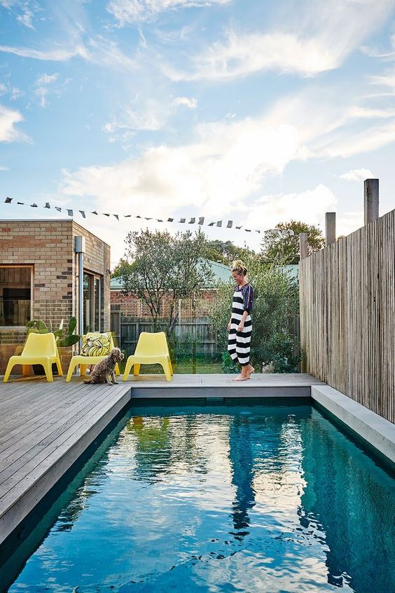 a lovely modern outdoor space with a wooden deck, a pool, bright yellow chairs, some greenery and cacti