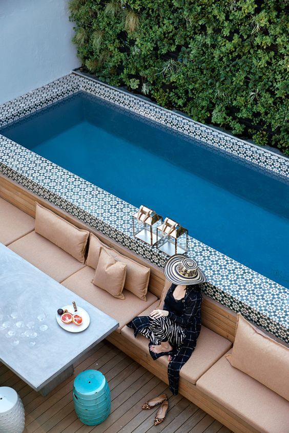 a lovely outdoor space with a wooden deck, a concrete table, a built-in bench with pillows and a narrow pool clad with bold tile