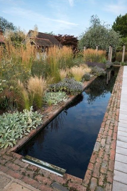 a lush and textured garden with bushes, grasses and leaves and a long and narrow pool clad with brick around that looks pretty natural