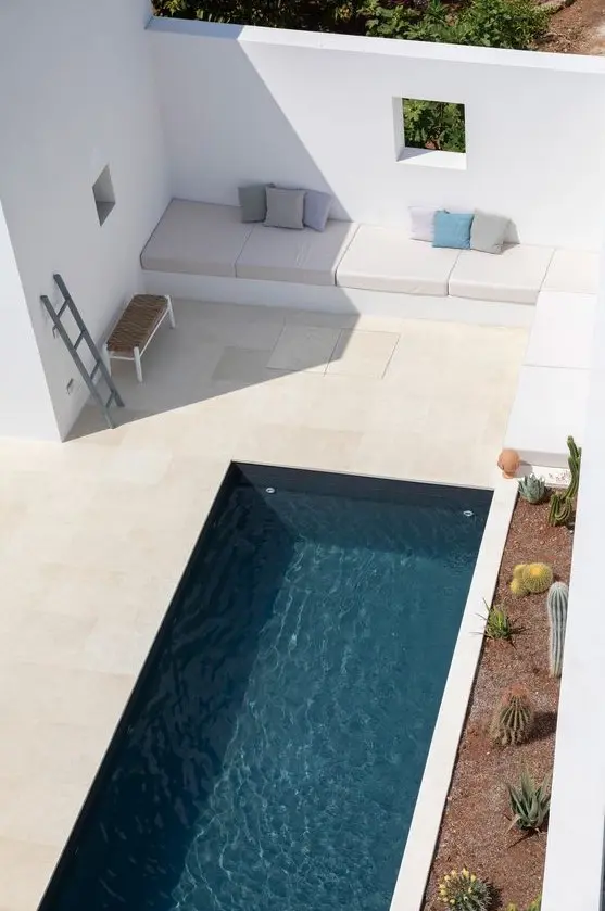 a minimalist outdoor space clad with tiles, with a long and narrow pool, a built in seat with pillows and some cacti