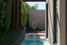 a minimalist outdoor space with a green wall and some beams over the space, a long and narrow pool and a tiled deck is all cool