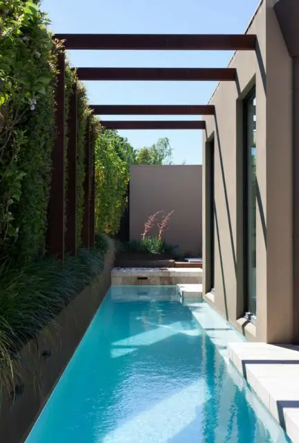 a minimalist outdoor space with a green wall and some beams over the space, a long and narrow pool and a tiled deck is all cool