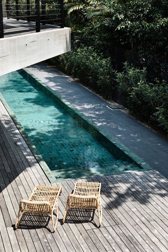 a minimalist outdoor space with a weathered wooden deck, a long and narrow pool and a couple of rattan loungers plus greenery around