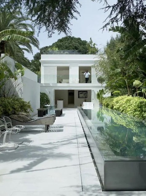a minimalist outdoor space with stone tiles, a long and narrow raised pool, loungers, chairs and greenery