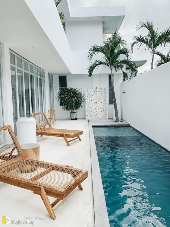 a minimalist white outdoor space with a long and narrow pool, woven daybeds, stools and stumps and potted trees