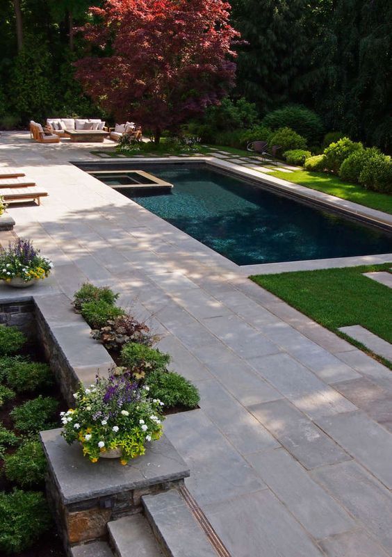 a modern outdoor space with a large stone deck and a pool, a green lawn and blooms and a sitting zone next to the pool