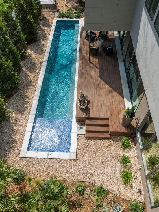 a modern outdoor space with trees, a wooden deck with potted plants, a long and narrow pool and some sitting zone
