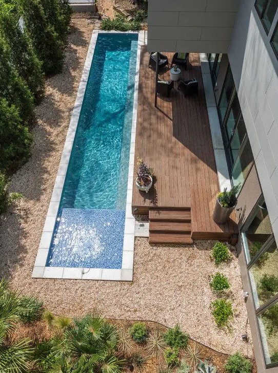 a narrow backyard pool clad with white tiles next to a wooden deck, with trees and greenery around