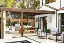 a neutral outdoor space with a pool, a white stone tile deck, greenery, chairs around a fire pit and a dining space under a roof