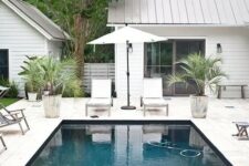 a neutral outdoor space with a pool, a whitewashed stone deck, neutral furniture, potted plants and some lights and lanterns