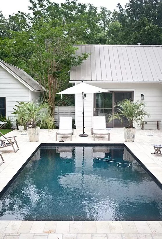 a neutral outdoor space with a pool, a whitewashed stone deck, neutral furniture, potted plants and some lights and lanterns