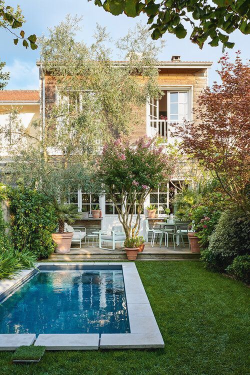 a pretty and welcoming backyard with a green lawn, a pool with a stone deck and a wooden one, outdoor furniture and potted plants and trees