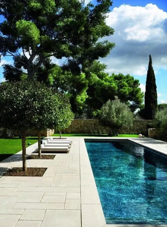 a refined outdoor space with a pool, a neutral stone deck, some loungers and some planted trees is a very cool space
