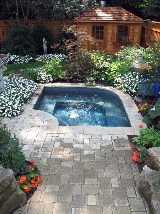 a small and lovely outdoor space with a small pool, a stone deck and lots of blooms around, with greenery and green lawn