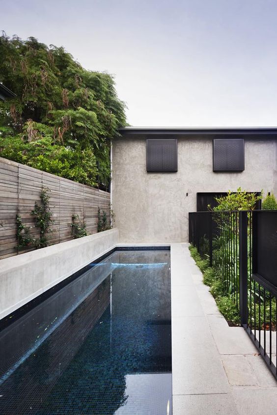 a small outdoor space with a stone deck, a long and narrow pool and some greenery plus a fence to keep it safe