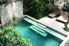 a small tropical backyard with a stone deck and loungers under the trees, a small pool and stone walls around to make it more private