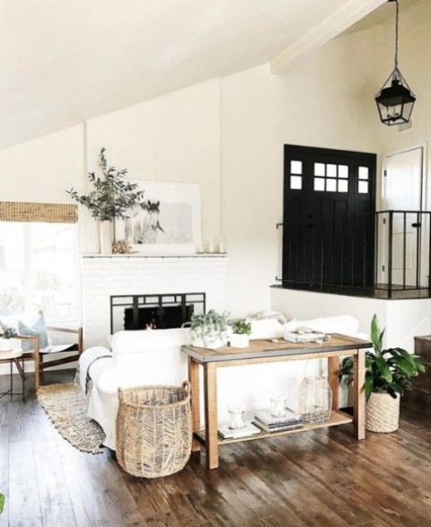a small white farmhouse living room with white furniture, wooden tables and chairs, baskets for storage and a brick fireplace