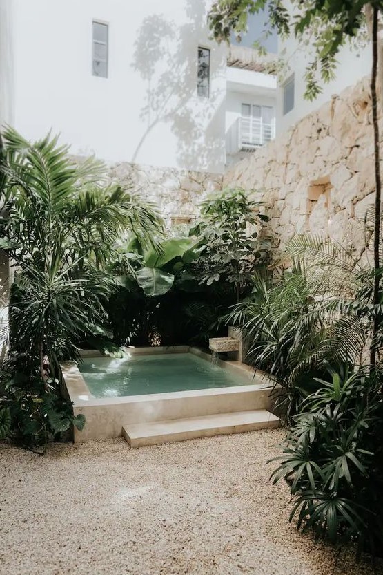 a tropical nook with stone walls, a plunge pool with a waterfall and stone borders, lots of tropical greenery around