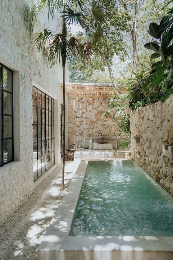 a tropical outdoor space with a stone fence, a narrow pool with a waterfall, a seat with pillows and some trees over the space