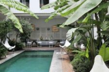 a welcoming outdoor tropical space with some white furniture, a brick deck, a long and narrow pool and some plants around