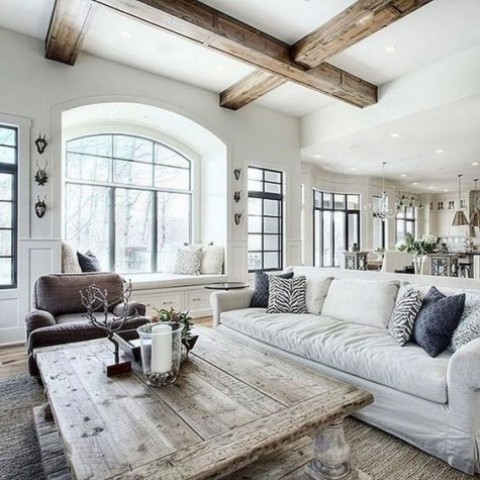 an airy farmhouse living room with a white sofa, a brown chair, a vintage whitewashed wooden coffee table and wooden beams