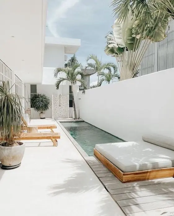 an all-white outdoor space with a deck, loungers, a daybed, a long and narrow pool and some potted trees is amazing