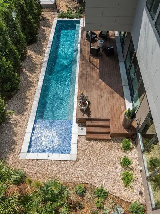 an outdoor space with trees, a wooden deck, a long and narrow pool, some growing plants and a sitting zone