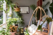 a boho porch turned into an orangery with lots of potted greenery and blooms and a large hanging rattan chair plus a rug