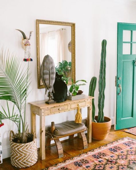 a bright boho entryway with a sleek wooden console, a catchy stool that matches, potted plants, a mirror in an arnate frame and some boho art