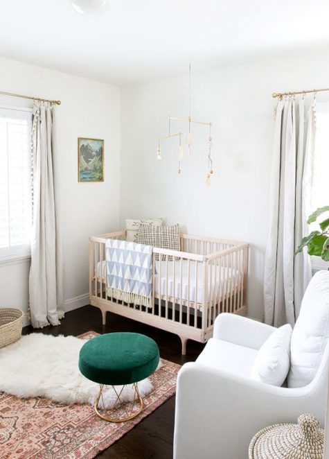 a chic gender neutral nursery in white, with a pretty mobile, a green ottoman, boho rugs and pompom curtains