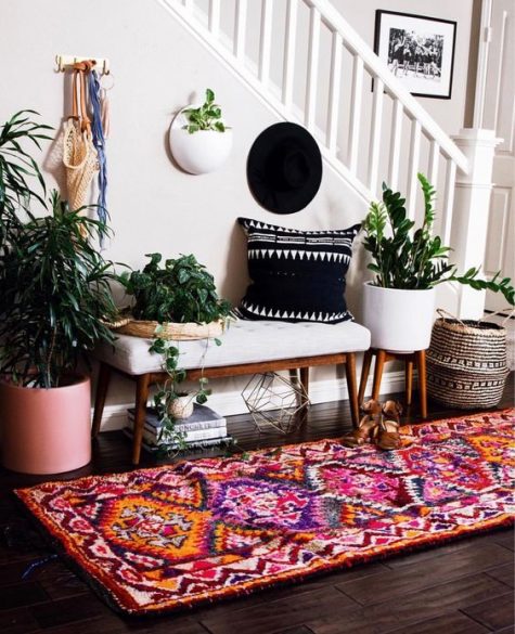 a colorful entryway with a white bench, a colorful boho rug, potted plants, a black printed pillow and baskets for storage