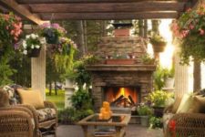 a cozy rustic terrace with a stone fireplace, wicker chairs asnd a table, lots of potted blooms and wooden beams