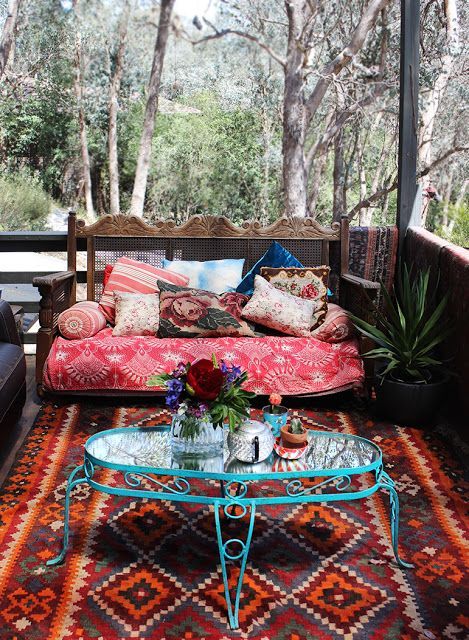 a gypsy meets vintage porch with bright boho textiles, lots of pillows, a blue forged coffee table and a carved wooden bench