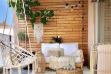 a neutral boho porch with wooden furniture, a rattan hanging chair, wooden lanterns, a jute ottoman, greenery and lights