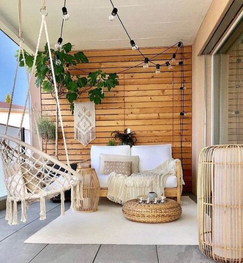 a neutral boho porch with wooden furniture, a rattan hanging chair, wooden lanterns, a jute ottoman, greenery and lights