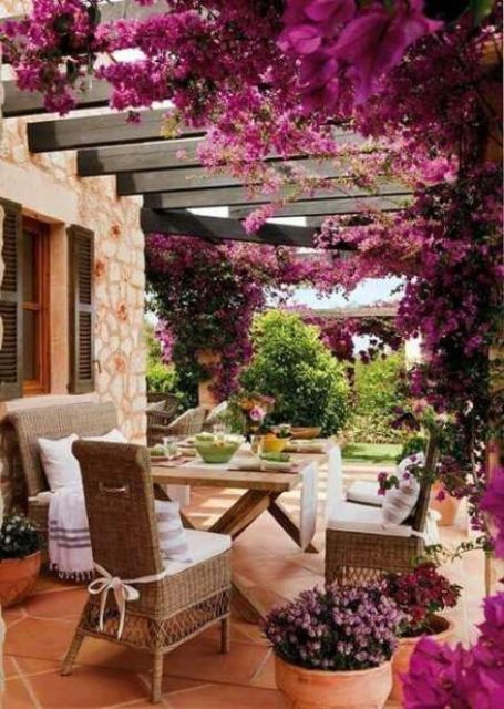 a rustic patio done with wicker furniture, a trestle wooden dining table, potted blooms and lots of wisteria