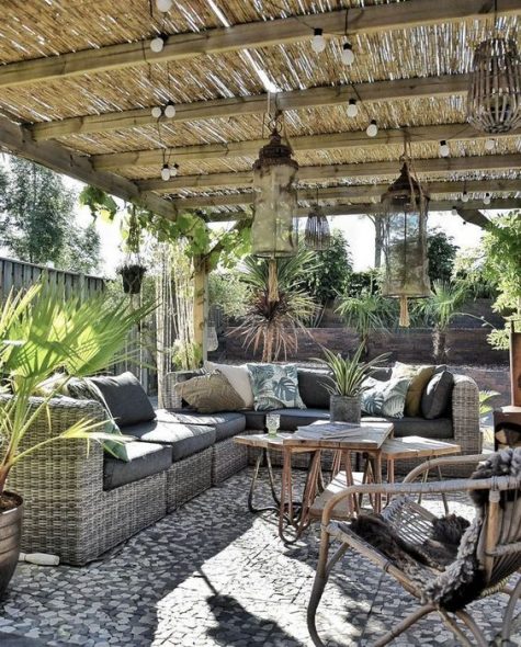 a small rustic patio with a large wicker sofa, rattan chairs and a table, lights and large lanterns plus greenery