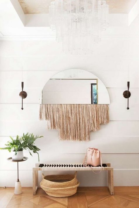 a whimsy boho entry with a woven bench, a basket for storage, a mirror with long fringe and a potted plant on a stand