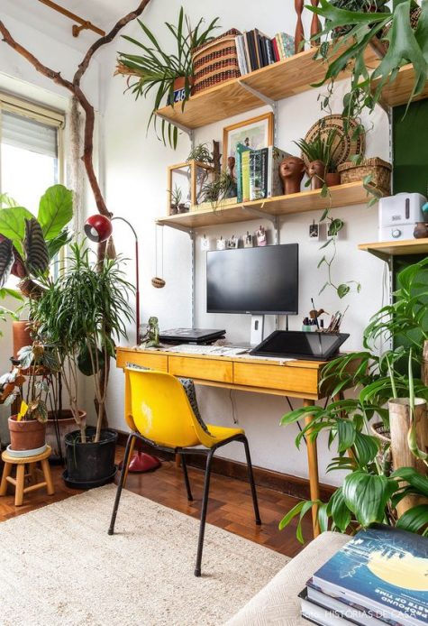 a bright boho home office with open shelving, a retro desk, a yellow chairs and lots of potted plants here and there