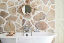 09 a light-filled farmhouse bathroom with a natural stone wall, a refined mirror and a clawfoot bathtub