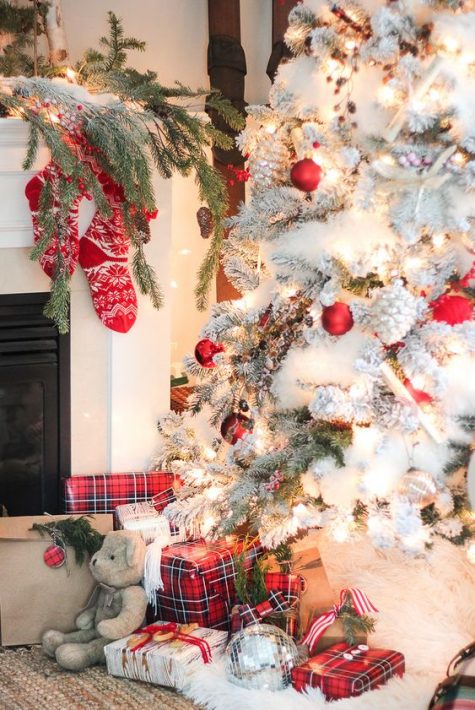 a gorgeous flocked Christmas tree with lights and red and white ornaments, plaid red gift boxes and red stockings by its side
