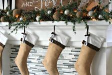 a rustic Christms mantel with a large sign, an evergreen and pinecone garland, lights, burlap balls and burlap stockings