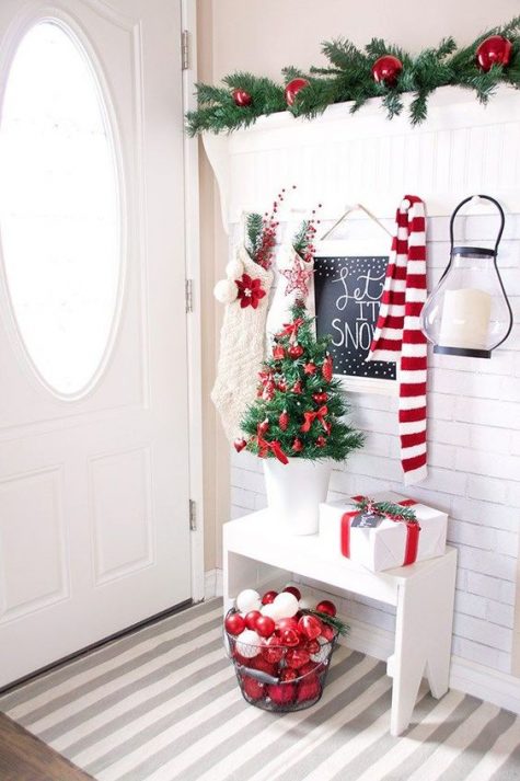a small and bright Christmas entryway with evergreens, white and red ornaments, a small tree, a scarf and stockings