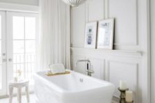 an elegant neutral bathroom with paneled walls, a pendant lamp, some white furniture and curtains