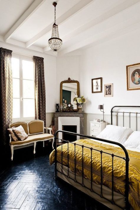 a beautiful Parisian bedroom with printed curtains, a crystal chandelier, a retro metal bed, a chic mustard loveseat and a marble clad fireplace