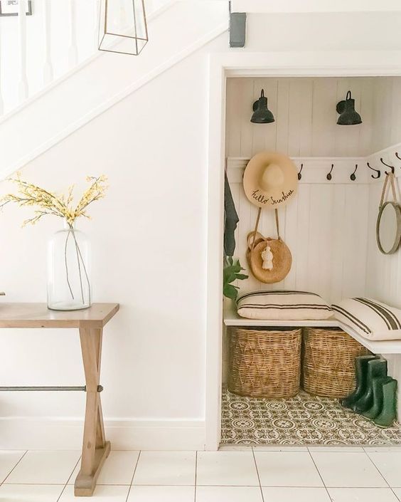 a small and cozy under stairs mudroom with a built-in bench, baskets and hooks, black sconces and some potted plants