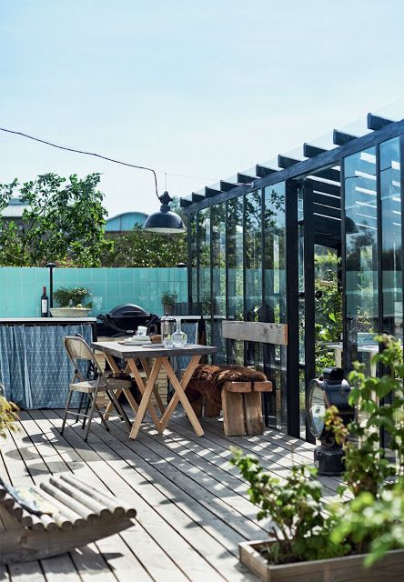 a Scandinavian terrace with a black greenhouse, potted greenery, wooden furniture, an outdoor kitchen and simple lamps