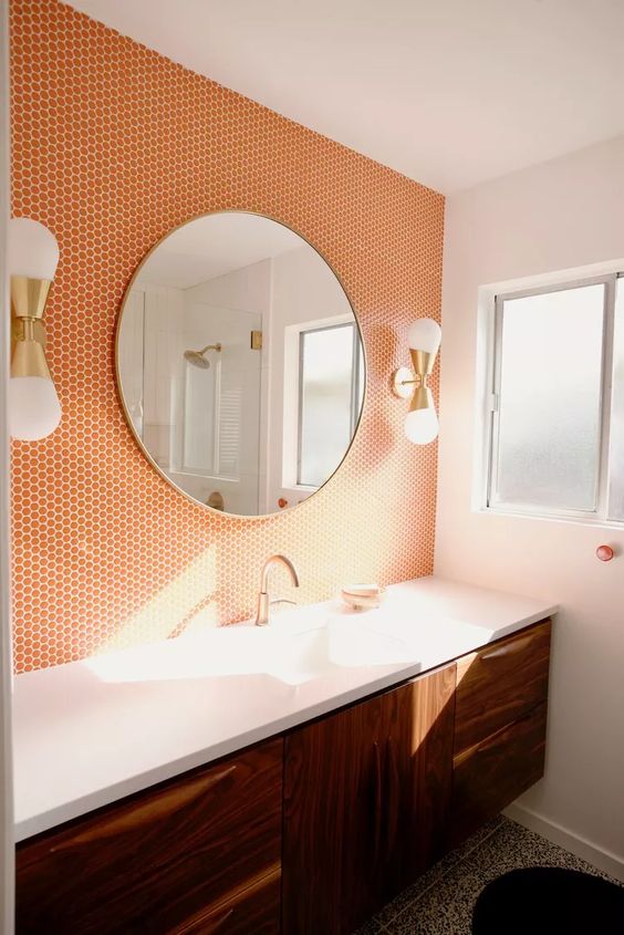 a bold sink space with orange penny tiles, a stained vanity, a round mirror, wall lamps is amazing